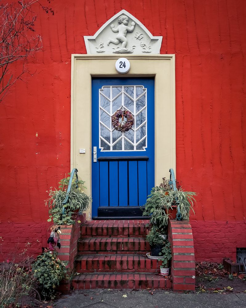 Caption: A photo of an entrance to a red building with brick steps and a bright, blue door with a floral wreath hung on it. There are potted plants on and around the steps. Above the door is the number “24” and a sculpture of a child running. (Local Guide @Potsdamomente)