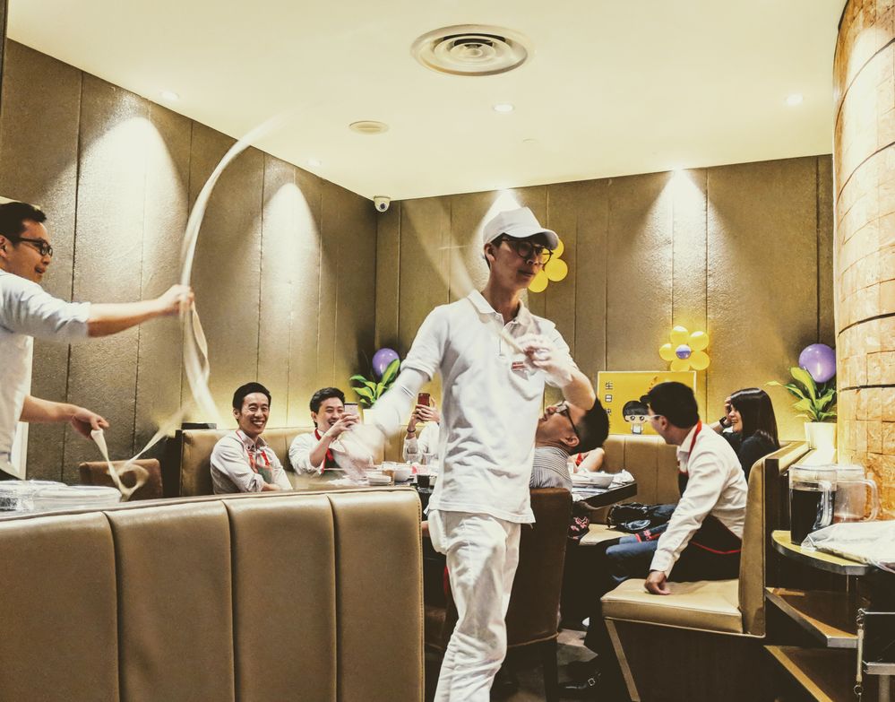 Caption: A photo of two members of the staff at Haidilao Hot Pot @313, dressed in white uniforms, dancing with a long thread of noodles in front of smiling customers. (Local Guide pianized)