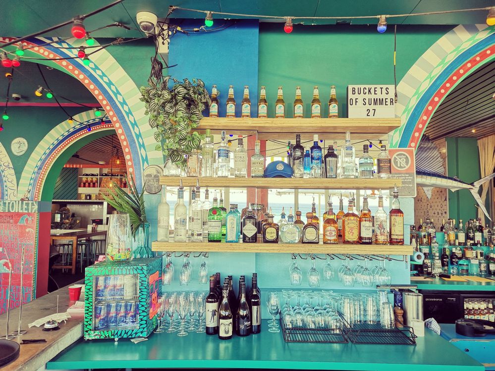 Caption: A photo of the turquoise bar at Sydney restaurant The Bucket List, showing rows of liquor and wine bottles on wooden shelves as well as glassware, bottles of beer, multi-colored string lights, and the restaurant’s colorful painted walls. (Local Guide Luigi Avantaggiato)