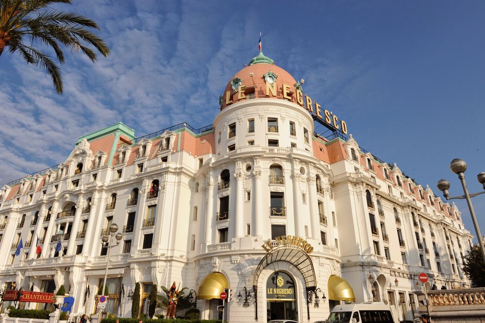 Caption: A photos of the Hotel Negresco in Nice, France showing it's cream, pink, and turquoise Art Deco exterior with large gold letters spelling out the hotel's name on the domed roof and smaller gold letters over the entrance to the lobby. (Local Guide Alberto Gozzelino)