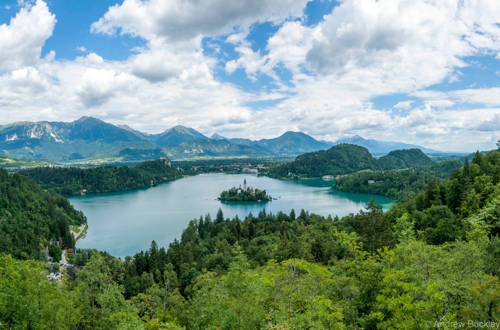 Caption: An aerial view of Lake Bled, Bled, Slovenia. Green trees and mountains surround the lake, and there’s a small island within it, with a church on top. (Local Guide @@Buckolujo)