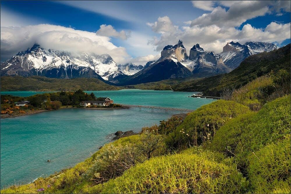 Caption: A photo of Torres del Paine National Park in the Magallanes Region of Chile showing mountains in the distance stretching up to the puffy, white clouds in the blue sky as well as a turquoise body of water with a long bridge leading to a building on a small island, all surrounded by greenery and hills. (Local Guide Fredy Costas)
