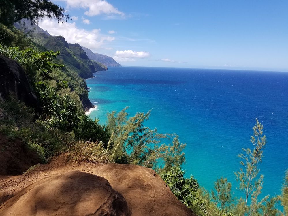 Caption: A photo from Kalalau Trail in Kauai County, Hawaii showing a cliff that's on the trail and the greenery that surrounds it as well as the turquoise and blue ocean below and mountains covered with greenery in the distance. (Local Guide Kyle Anderson)