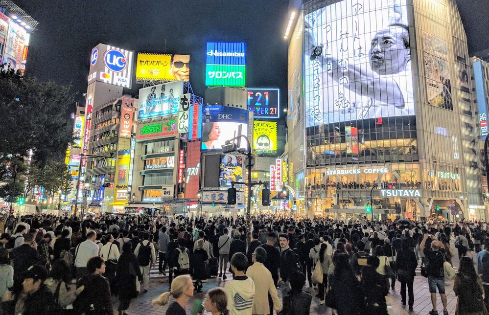 Caption: A photo of a large crowd of people walking in the Shibuya Crossing with illuminated advertisements on buildings in the background. (Local Guide Jorge Pinto)