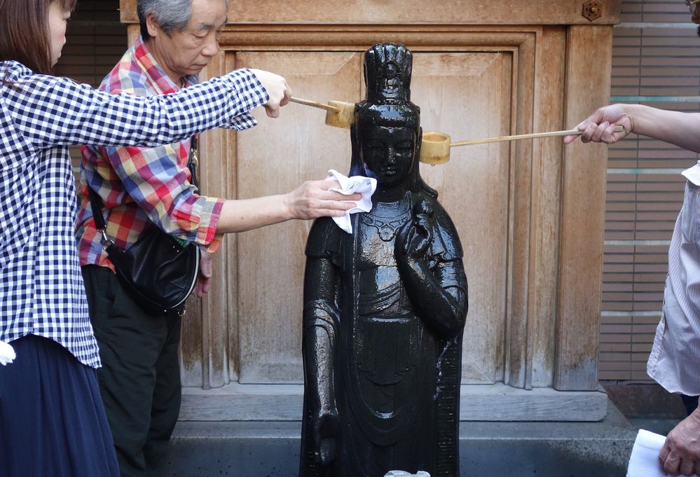 Caption: A photo of three people wetting and polishing Togenuki Jizo, splinter-removing Buddha statue located along Sugamo Street in Tokyo, Japan. (Local Guide Ken Kurasawa)