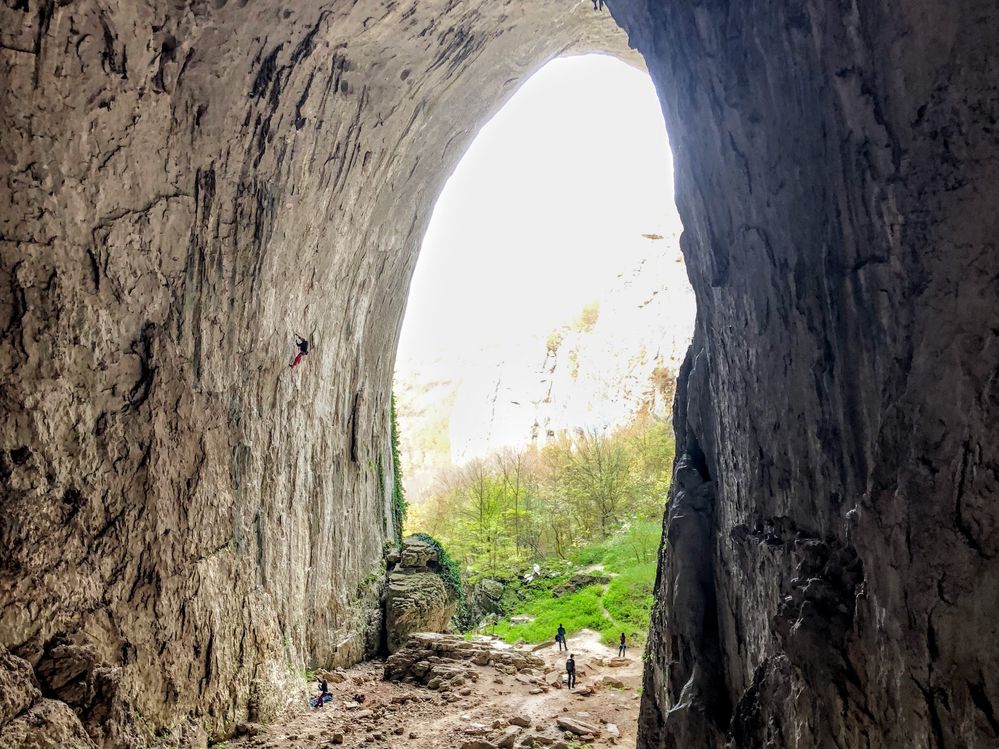 Caption: A photo of the exit of Prohodna cave, with a person climbing on one of the very tall walls, and three people going out in a green meadow behind the cave. (Local Guide @sonnyNg)