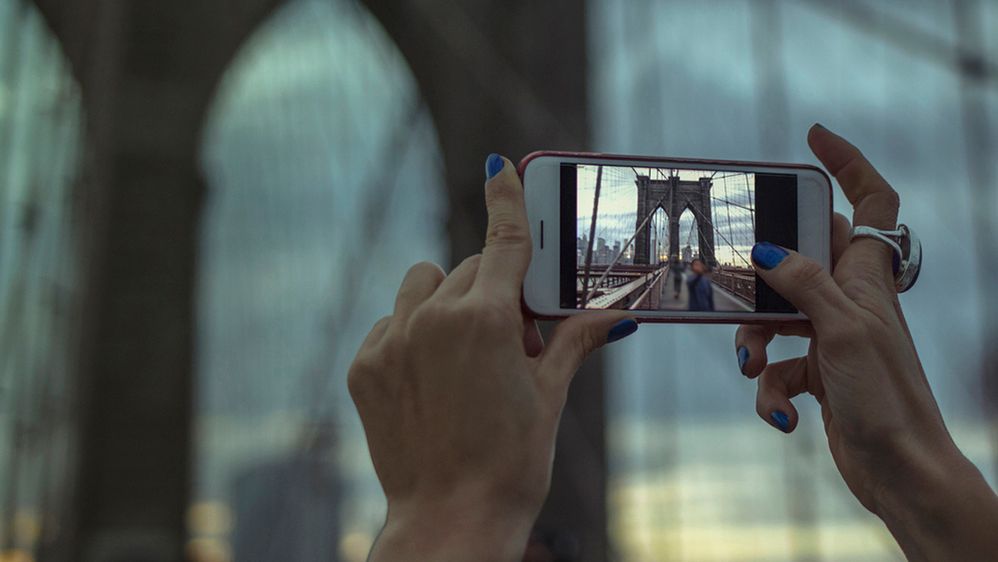 Caption: A closeup photo of a woman’s hands holding a smartphone and taking a photograph of the Brooklyn Bridge at dusk. (Getty Images)