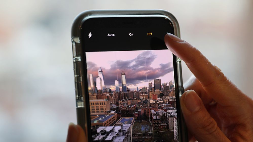 Caption: A closeup photo of a woman’s finger turning off the flash on a smartphone, with a view of New York City during twilight in the camera frame. (Local Guide Christina-NYC)