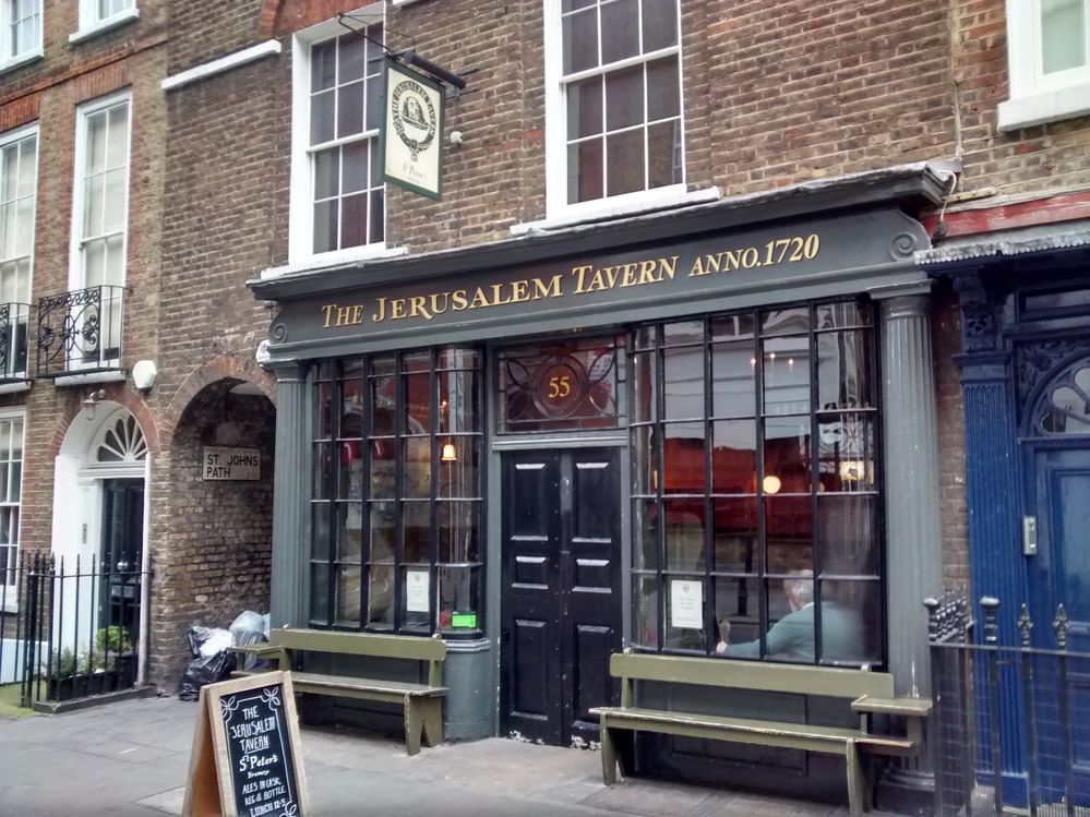 Caption: A photo of the brick and gray exterior of The Jerusalem Tavern in London showing its two large front windows, gold nameplate over the door, and green benches out front. (Local Guide Matthew Bradby)