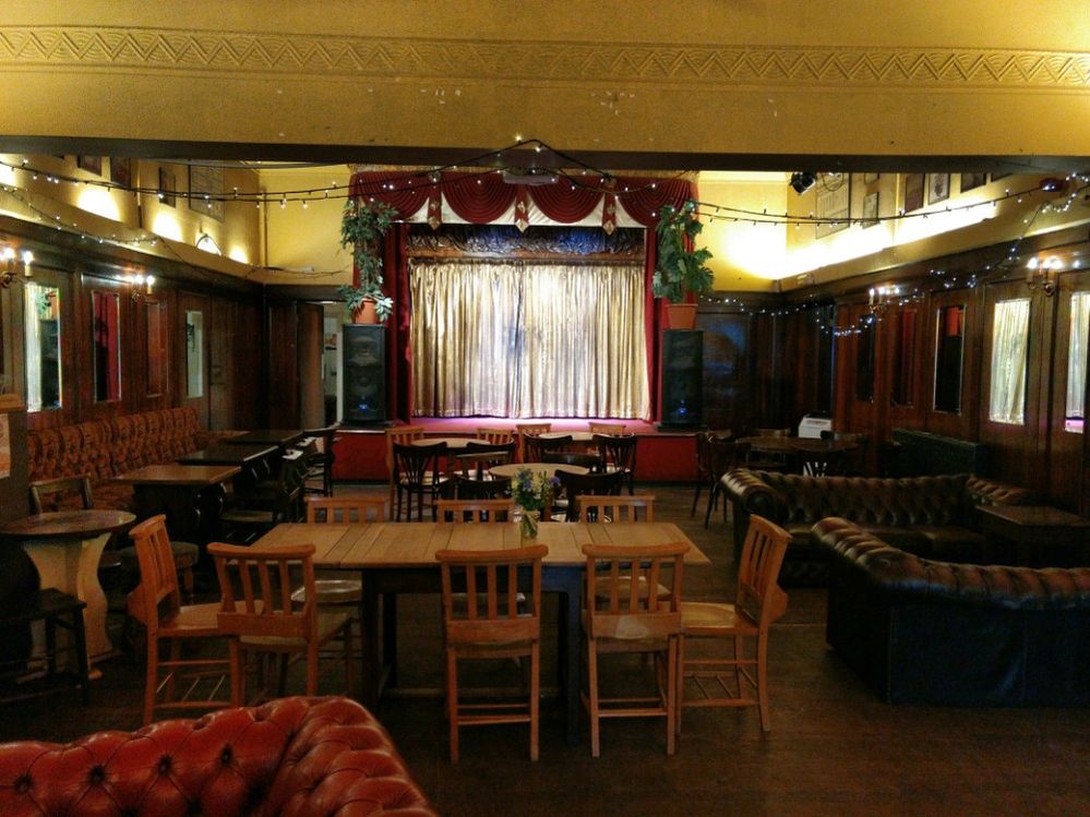 Caption: A photo of the interior of The Ivy House, showing wooden tables and chairs, leather sofas, twinkle lights strung from the ceiling, and the stage draped with red and gold fabric. (Local Guide J Mark Dodds)
