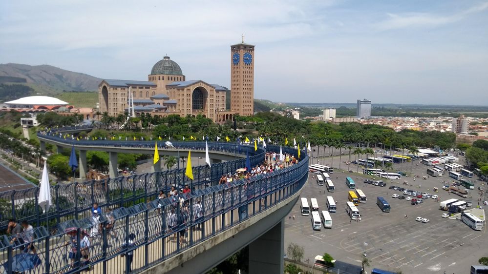 Caption: A photo of the Walkway of Faith, a path above the ground, leading to the Basilica of Our Lady Aparecida (in the distance), with a lot of pilgrims walking on it. (Getty Images)