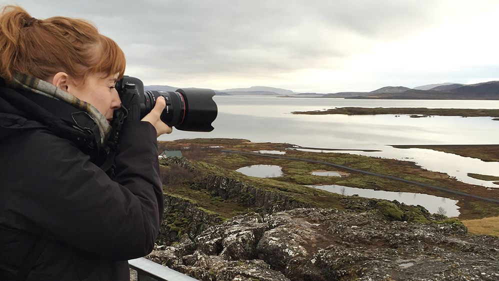 Caption: A photo of Local Guide Christina-NYC taking a photograph with a camera, overlooking a lake and hills in Iceland. (P. Hegarty)