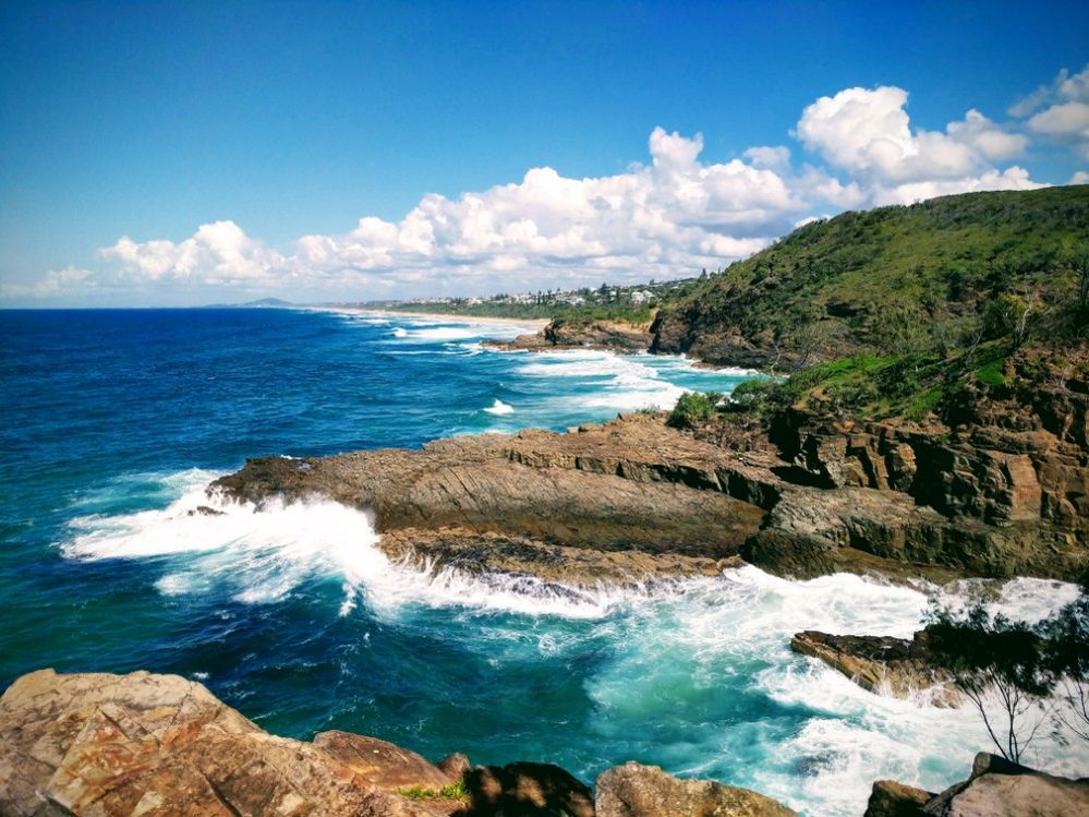 Caption: A photo of Noosa National Park in Queensland, Australia, showing the coastline with beautiful view of the blue and turquoise ocean, cliffs jutting out into the waves, and lush hilltops. (Local Guide Chris Bogin)