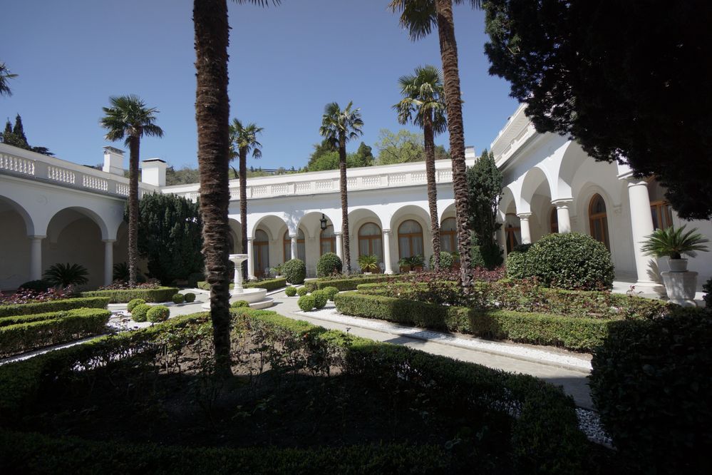 Надпись: Фотография внутреннего Итальянского сада Ливадийского дворца. Белый внешний коридор окружает зеленые кусты и высокие пальмы. (Местный Эксперт @MashaPS)