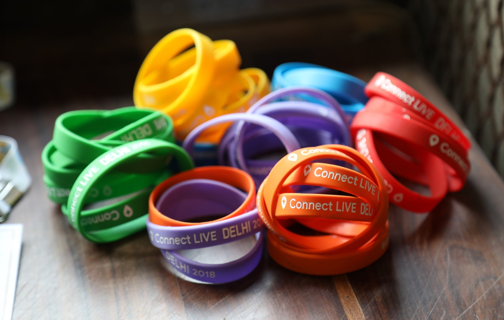 Caption: A photo of red, orange, purple, green, yellow, and blue wristbands that say “Connect Live DELHI 2018” placed on a wooden table.