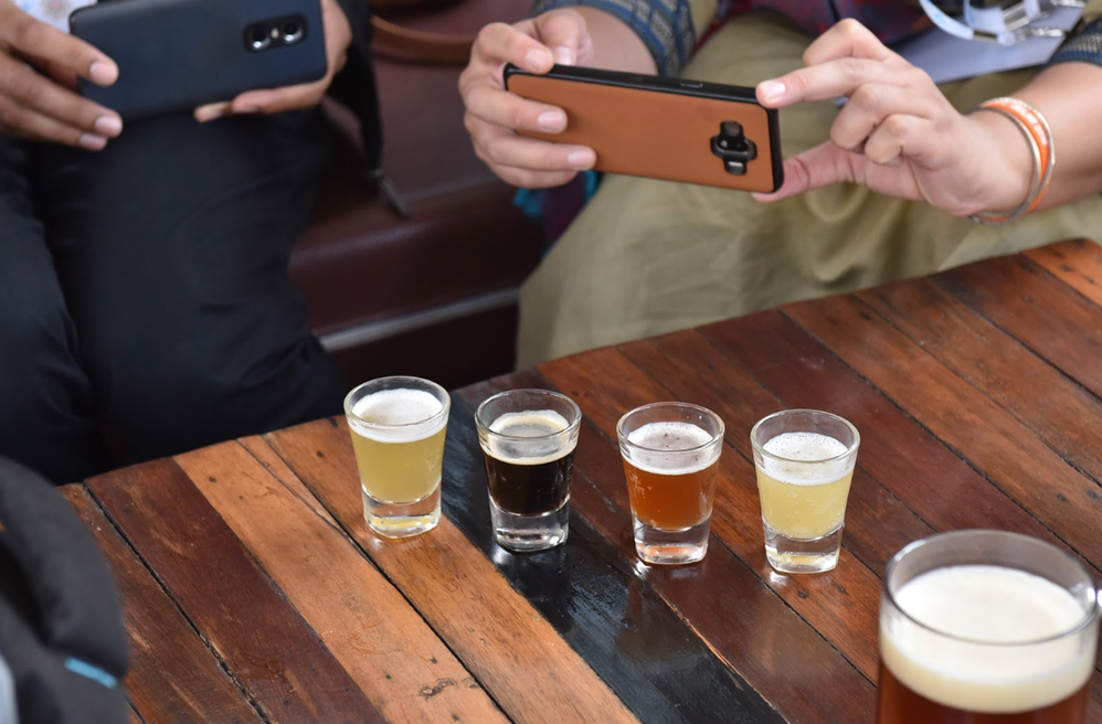 Caption: A photo of two people taking a photo of four shot glasses filled with different kinds of beer using their smartphones at Connect Live Delhi 2018.