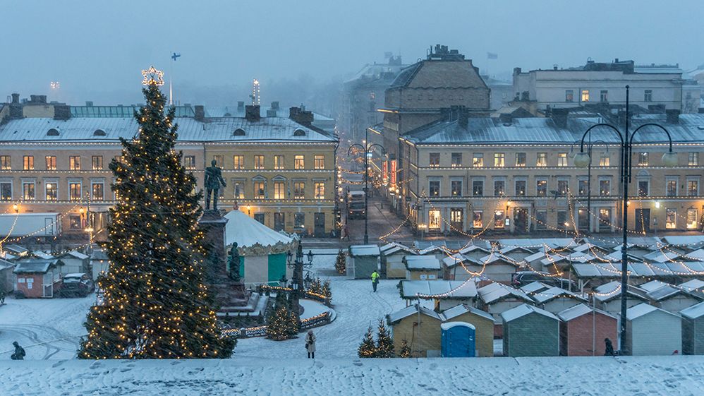 Caption: A photo of the Senate Square in Helsinki, with a tall Christmas tree covered in lights, and many colorful stalls, built for the Christmas market. Everything is covered in snow, and there are lights hanging over the stalls. (Getty Images)