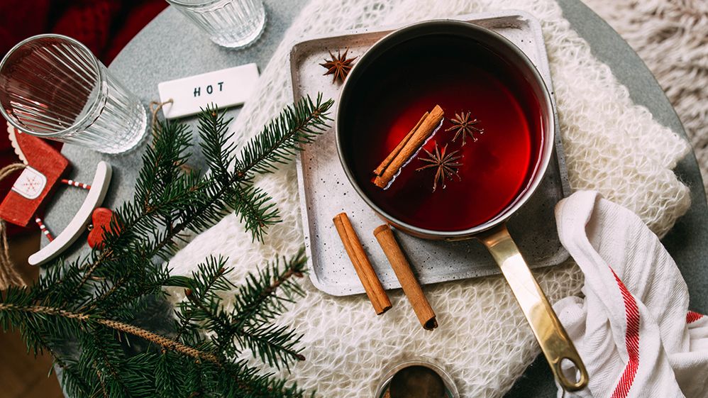 Caption: A photo of a tin pot with mulled red wine, a cinnamon stick, and two star-shaped spices on a tray. Artistically arranged around it are a towel, a green fir tree branch, a Christmas toy, two water glasses, two cinnamon sticks, and a tile with the word “hot” written on it. (Getty Images)