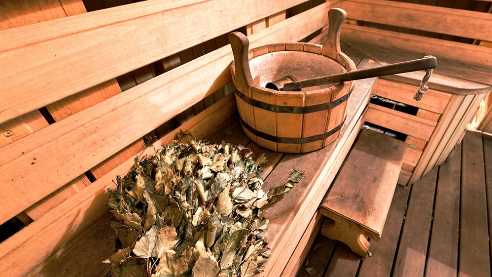 Caption: A photo of a wooden bench inside a sauna, with a birch branch with leaves and an empty bucket with a ladle inside of it. (Getty Images)