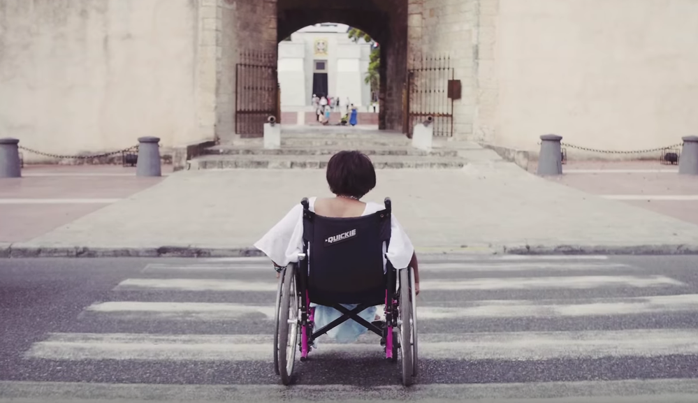 Caption: A screenshot from a Local Guides YouTube video called “Local Hero: Mapping For Good in the Dominican Republic,” showing a woman using a wheelchair crossing a street at a crosswalk.