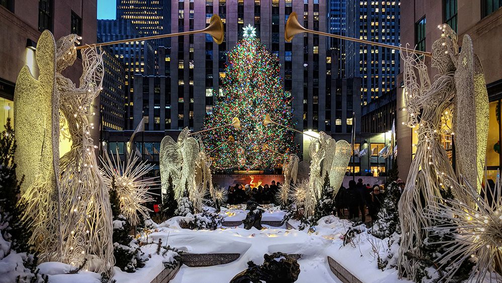 Caption: A photo of a snow-covered holiday display at Rockefeller Center in New York, NY, which includes a Christmas tree and statues of trumpeting angels. (Local Guide Jonas Modesto)