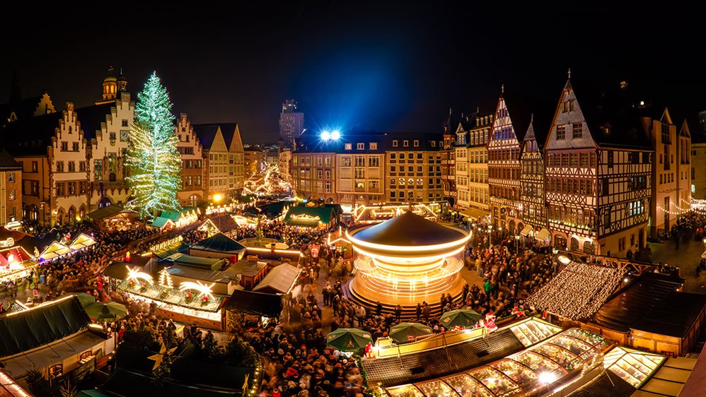 Caption: A photo of a night time German Christmas market taken from a high point. There are many people walking around different stalls, an amusement ride, and a Christmas tree, all brightly decorated, with Bavarian-style buildings surrounding the square. (Getty images)