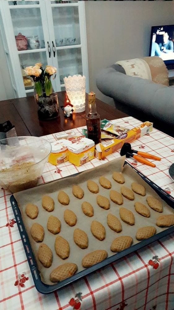 Caption: A photo of a baking tray full of melomakarona sweets, ready to be baked, on a white and red tablecloth. On the table, there are also a bowl of cookie dough, two packages of flour, a bottle, a package of parchment paper, a vase with flowers, a lamp, and an elf figurine. (Local Guide @VasT)