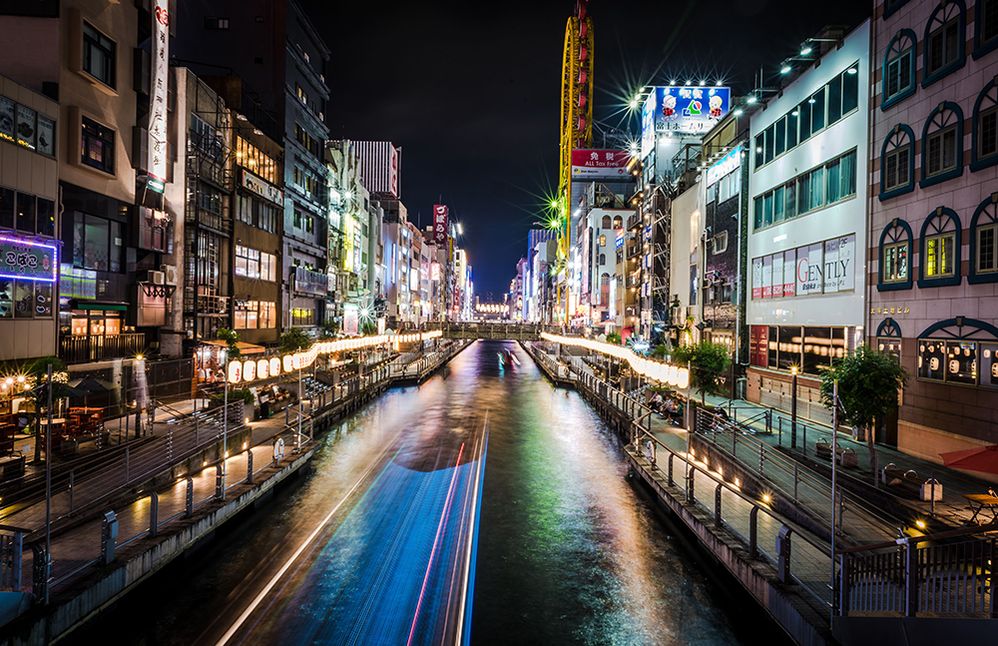 Caption: A photo of buildings lining the Dōtonbori canal at night, lit with neon lights. (Local Guide Kin ho)