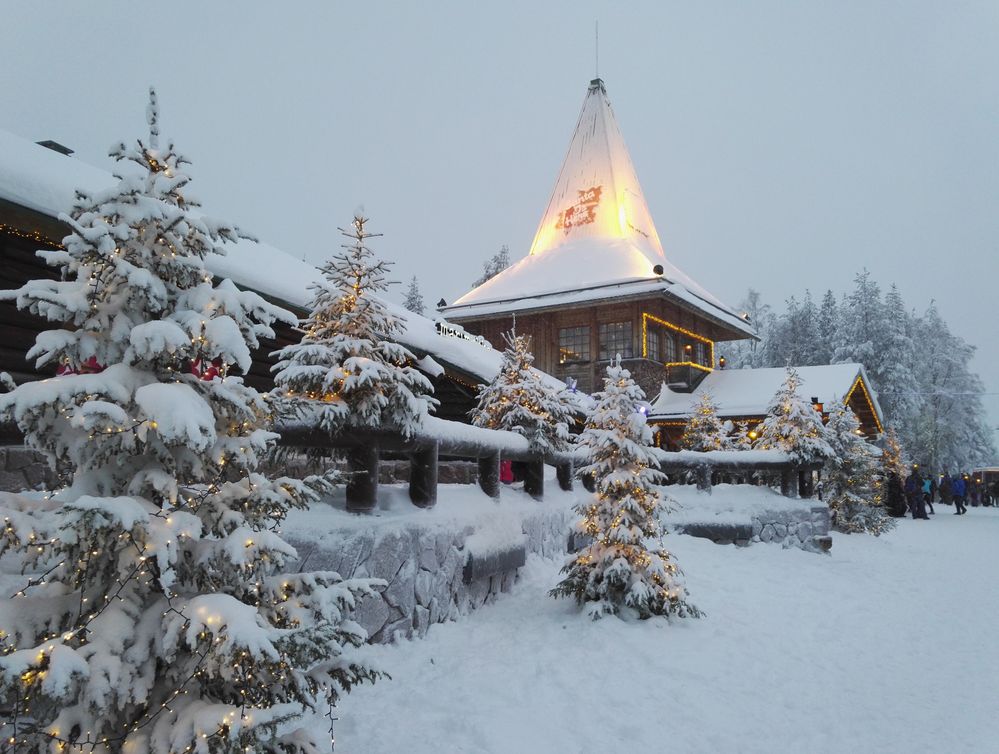 Caption: An exterior photo of a snowy wood chalet with Christmas trees decorated in white lights at Santa Claus Village in Rovaniemi, Finland. (Photo by Local Guide Ville Karhusaari)