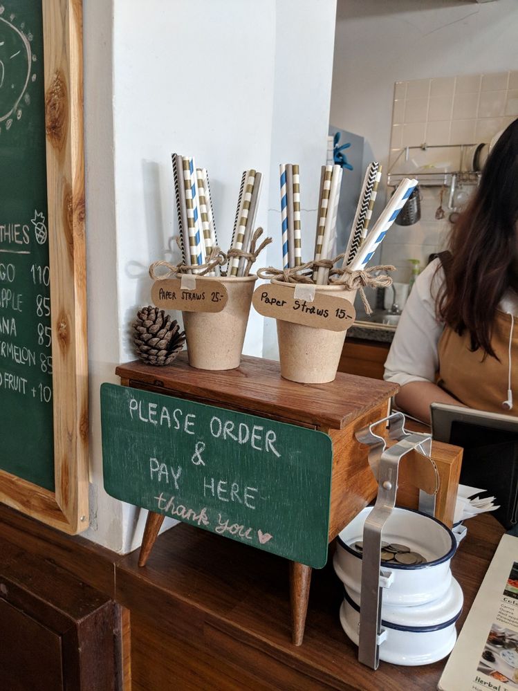 Caption: A photo of two buckets filled with paper straws placed on top of a wooden box with a sign over it that says, “Please order & pay here. Thank you,” at Better Moon cafe in Bangkok, Thailand. (Local Guide Suzanne Nam)