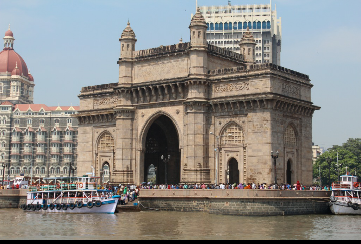 Caption: A photo of the Gateway of India in Mumbai, a large stone archway built in 1924, showing visitors crowded around the monument as well as the waterfront and boats in front it. (Local Guide Akshay Kongle)