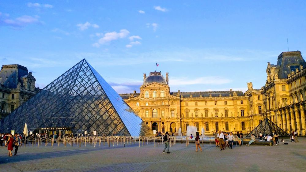 Caption: A photo of the exterior of the Louvre Museum in Paris, France, showing the beautiful Louvre Palace, fountain, one of the small glass pyramids, and the large glass pyramid well as the ropes designating the entry queue. (Local Guide Hanh Dung Le Vu)