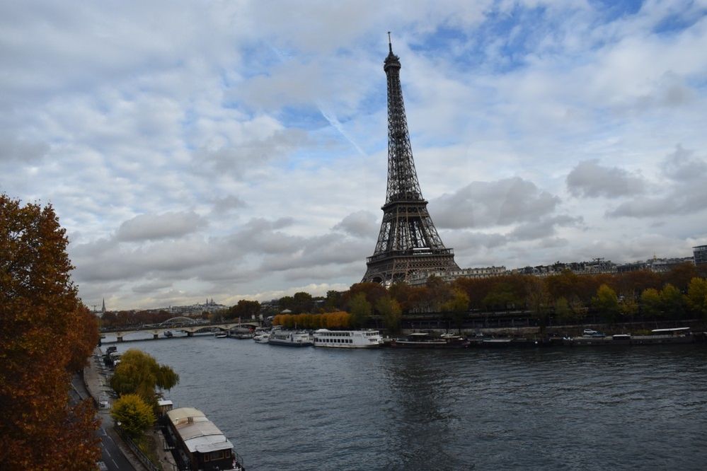 Caption: A photo of the Eiffel Tower on a cloudy autumn day taken from the opposite bank of the Seine river in Paris, showing the tall wrought-iron lattice structure against the blue sky and white clouds. (Local Guide Sara Cuesta)
