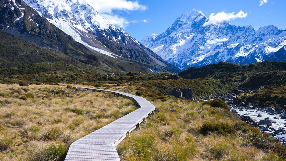 Caption: A photo of a wooden walkway in Aoraki/Mount Cook National Park in Canterbury, New Zealand, with a mountain range in the background. (Local Guide Will Warin Ratchananusorn)