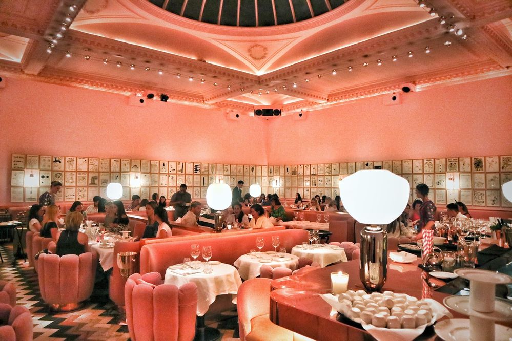 Caption: A photo of the interior of the London restaurant Sketch, showing its pink velvet chairs, banquets, and walls, as well as tables set with wine glasses and the bar. (Local Guide Cat Morley)