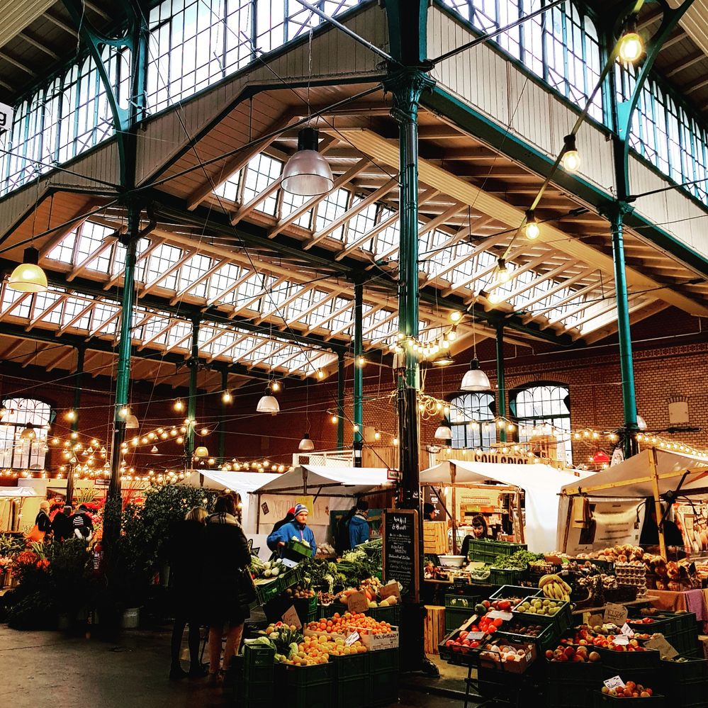 Caption: A photo of the inside of Markthalle Neun, an indoor bazaar in Kreuzberg, Berlin. There are big windows on the roof and yellow lights hanging over various food stands. (Local Guide @NadyaPN)