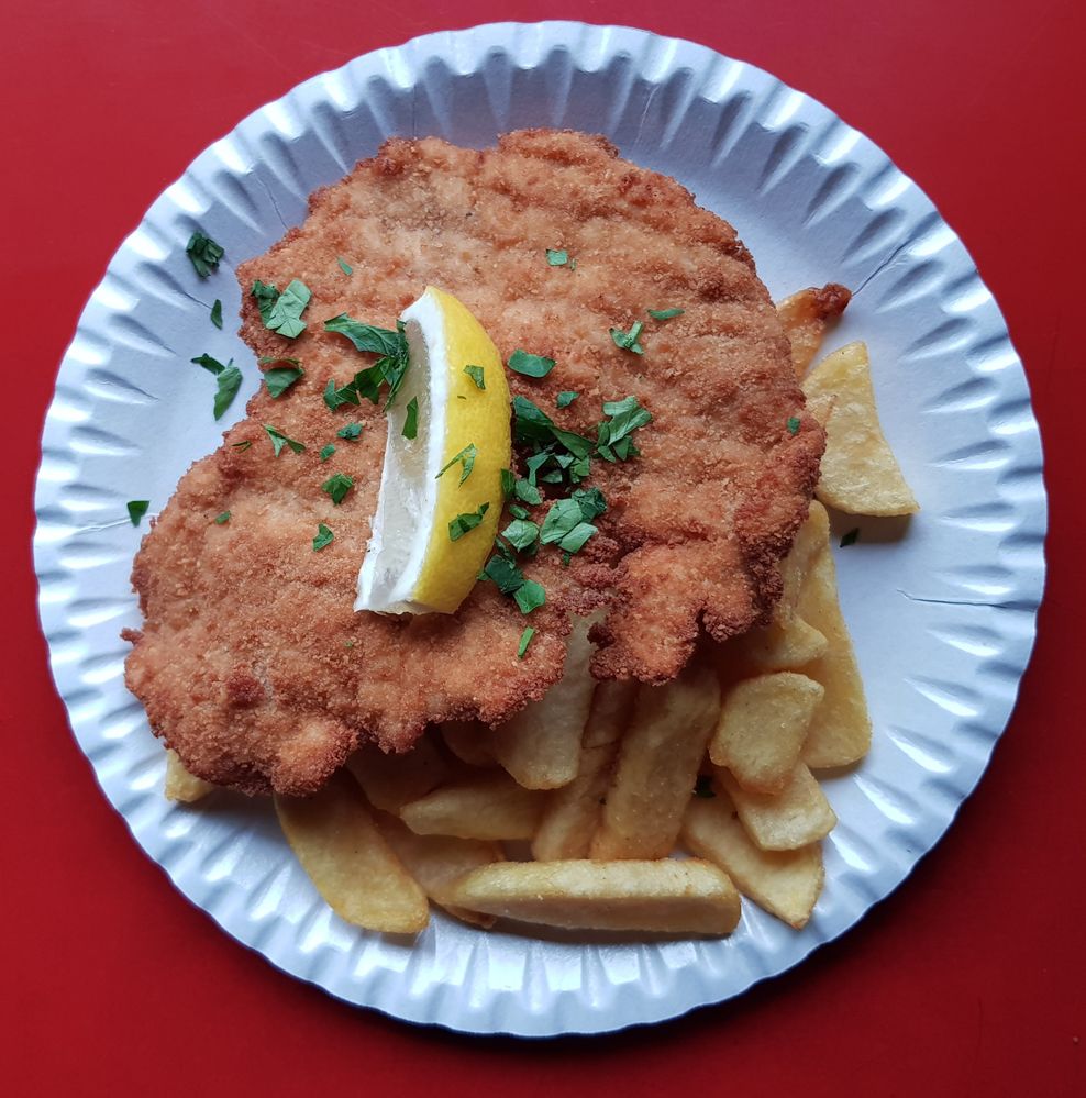 Caption: A photo of a paper plate with a schnitzel, fries, a slice of lemon, and parsley on a red table. (Local Guide @NadyaPN)