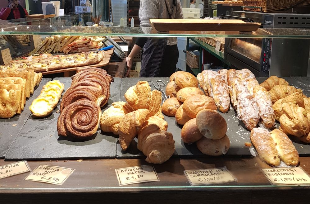 Caption: A photo of baked breads and pastries on display at a breakfast stall in Markthalle Neun, Berlin. There are croissants, brioches, muffins, slices of pizza, and more. (Local Guide @NadyaPN)