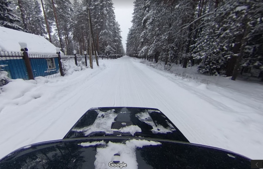 Caption: A screenshot of a Street View image that shows a black car driving down a snow-covered road surrounded by trees and a blue house behind a fence on the left in Kholuy, Russia. (Local Guide @METR)