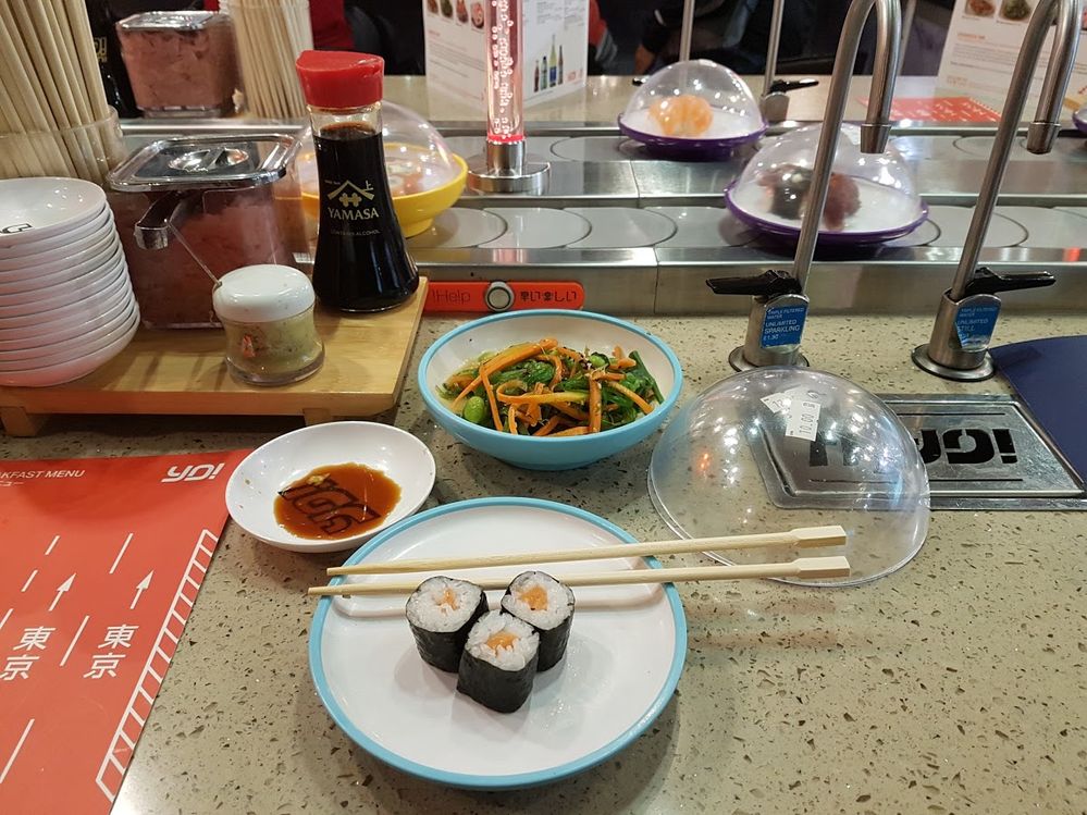 Caption: A photo of a plate of sushi and a bowl of vegetables on a counter, next to a conveyor belt of covered plates of food at Yo! Sushi at Heathrow Airport in London, UK. (Local Guide Amol Sinha)