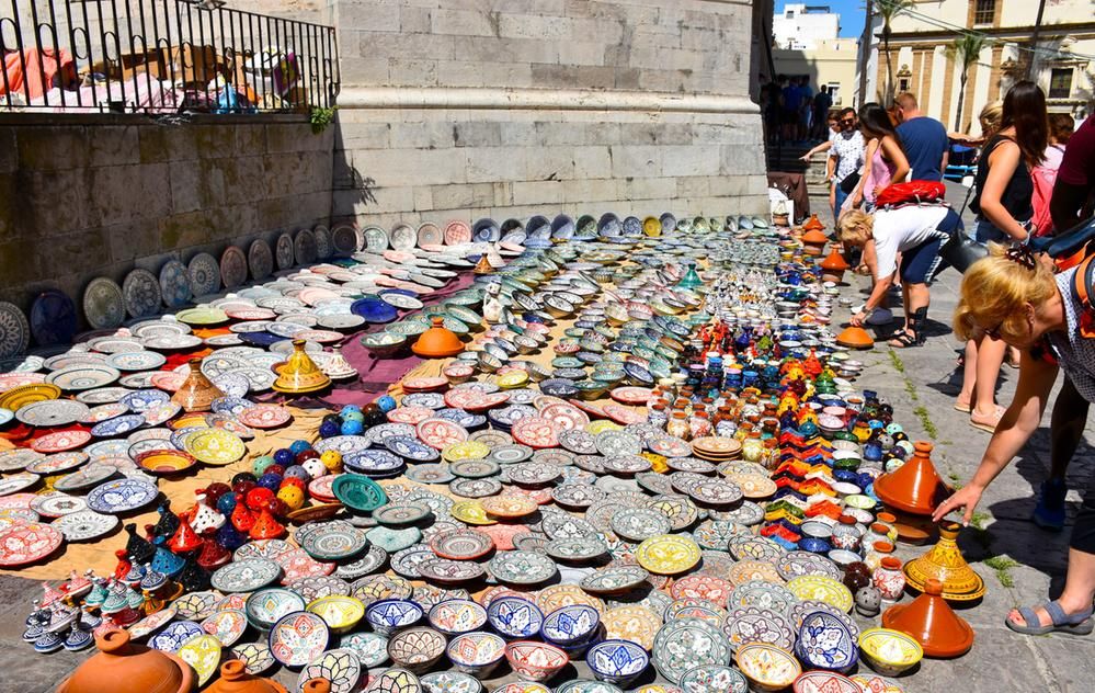Caption: A photo of tons of colorful and intricately designed dishes laid out for sale on the sidewalk in Cadiz, Spain, as people look and reach down to touch them. (Local Guide @Madina_Travelmania)