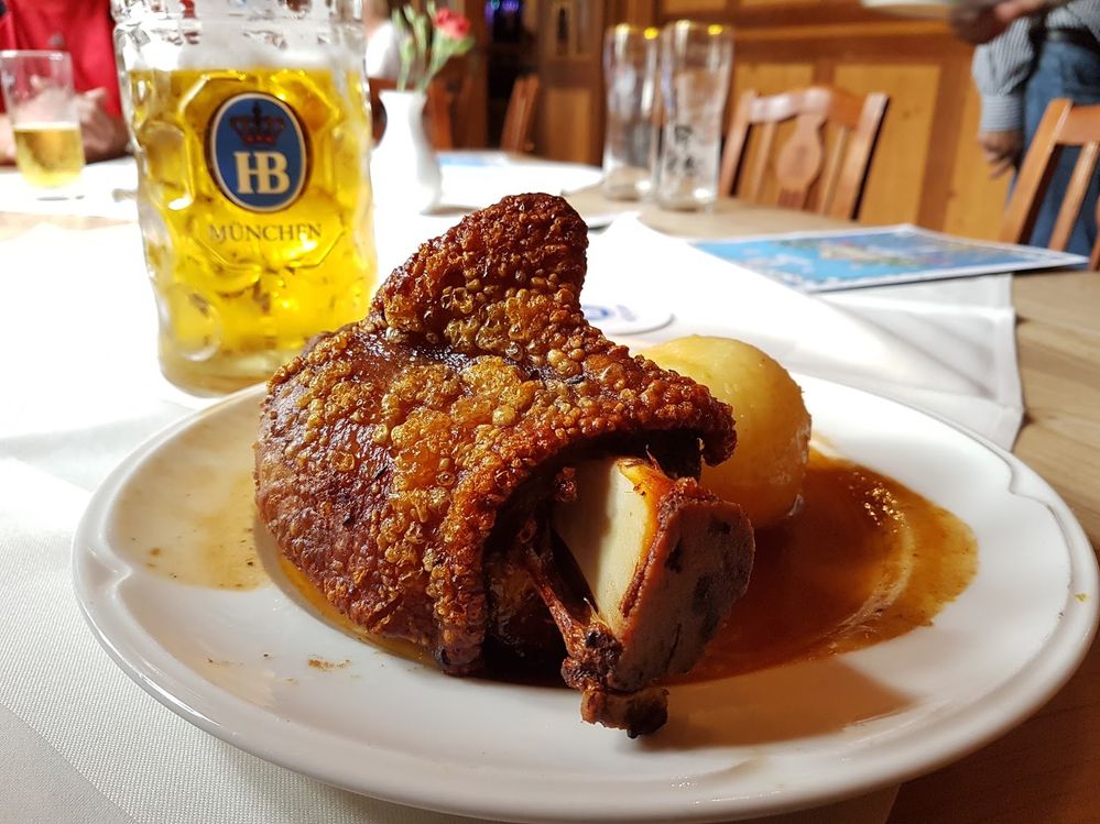 Caption: A close-up photo of a roast pork on the bone with crispy crackling and a potato dumpling sitting in a pool of gravy with a pint of beer in the background. (Local Guide Jeremy Banzhaf)