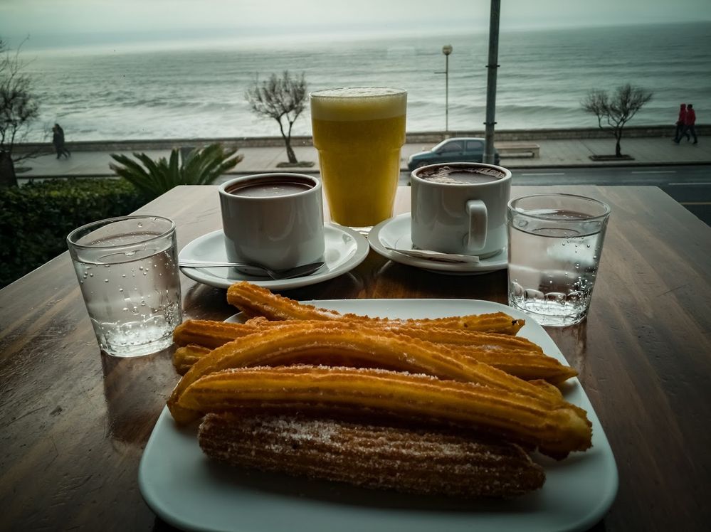 Caption: A photo of a plate of churros along with two glasses of water, two cups of coffee, and a glass of orange juice on a table next to a window that overlooks the beach at Manolo in Buenos Aires  (Local Guide Juan Carlos Lizarazu)