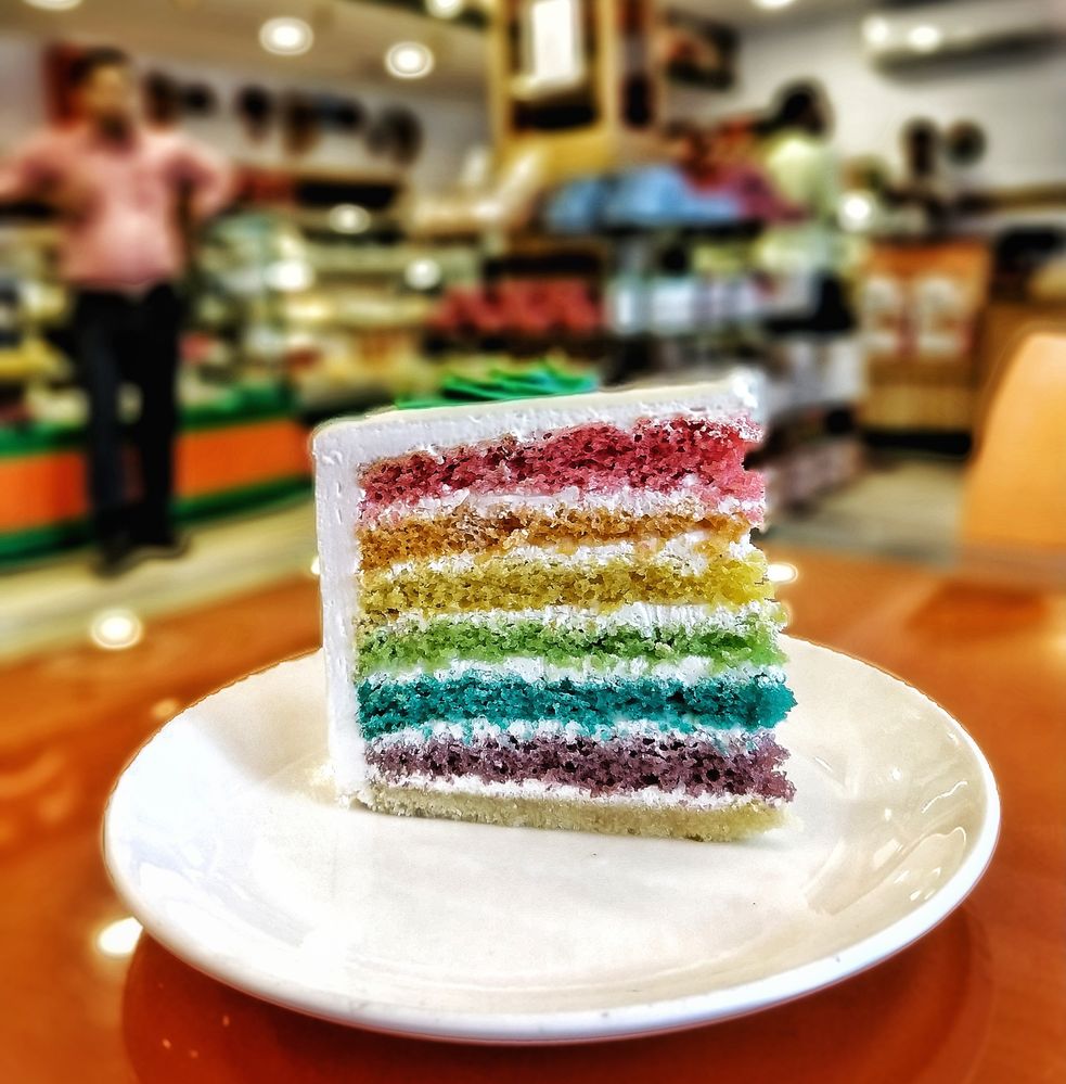 Caption: A photo of a slice of rainbow cake on a white plate at Bread World in Kerala, India. (Local Guide ananthu sajeev)