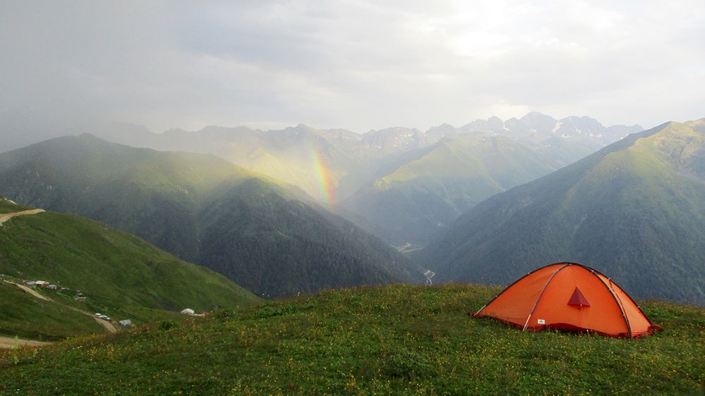 Caption: A photo of an orange tent on the top of a mountain with a view of a rainbow and mountain range beyond it. (Local Guide Kadir Erdogan)
