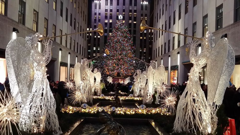 Caption: A photo of holiday decorations at Rockefeller Center in NYC including trumpeting angels and the famous Christmas tree lit with colorful lights, taken at night. (Local Guide to战乡生by Li)