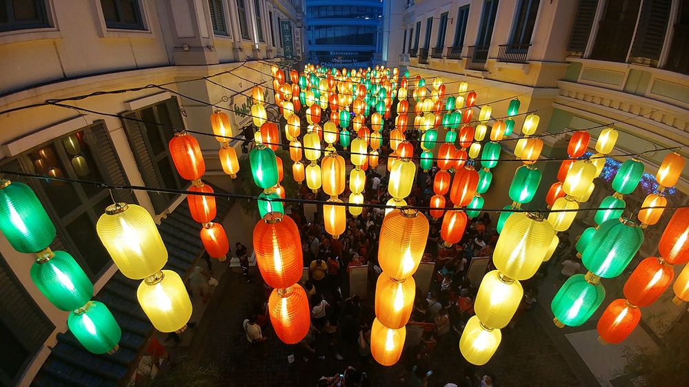 Caption: A photo of red, green, yellow, and orange glowing lanterns strung across a courtyard taken at night during Lunar New Year in Manila, Philippines. (Local Guide Richie See)