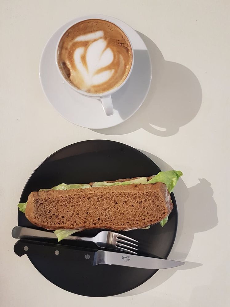 Caption: A photo of vegetarian sandwich with olive paste, lettuce and gouda cheese in a black plate. As well a cappuccino. (Local Guide @InaS)