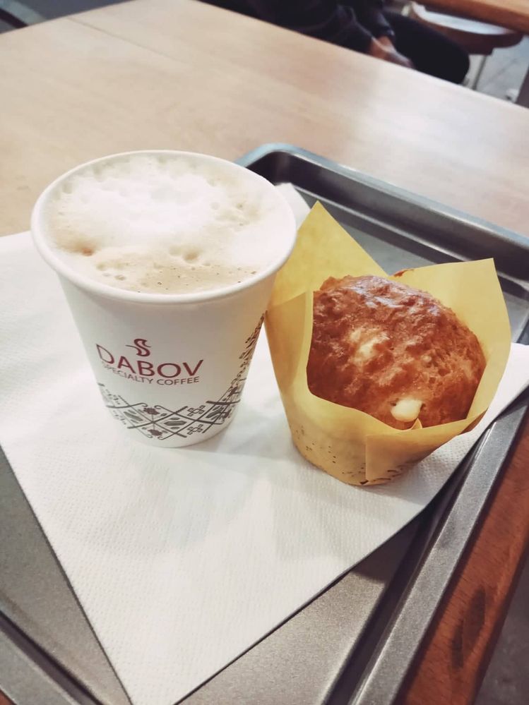 Caption: A photo of a tray on top of it there is a biodegradable cup of cappuccino and next to it a cheese muffin.