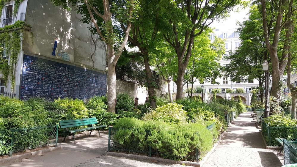 Caption: A photo of the Wall of Love in Paris, a mural made up of 612 tiles blue tiles with “I love you” written on them in many different languages in white paint, surrounded by trees and green plants. (Local Guide S N)